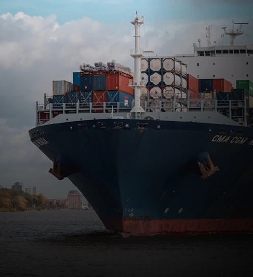Um grande navio de carga envolvido em transporte marítimo de cargas navega em um rio, carregado com contêineres coloridos, enquanto árvores e prédios distantes se alinham na costa sob um céu nublado.