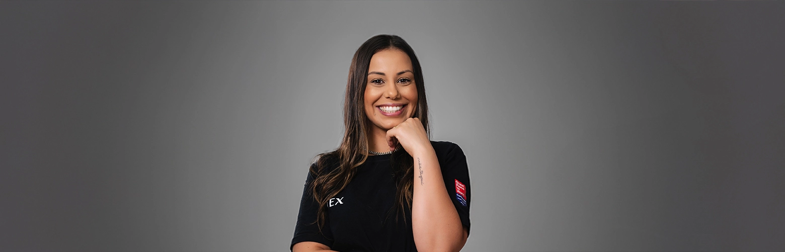 Uma mulher de cabelos castanhos longos está em pé em frente a um fundo cinza liso, sorrindo e vestindo uma camiseta preta com texto em amarelo e branco com palavras relacionadas a transporte, logística e agenciamento de cargas.