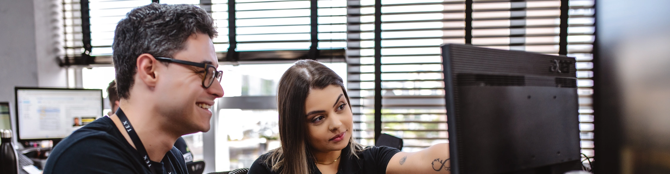 Homem e mulher da equipe TEX colaboram em frente a um computador; ele sorri enquanto ela aponta para a tela com expressão atenta. Ambiente de escritório com persianas ao fundo.