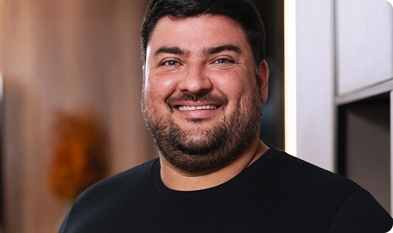 Um homem com cabelos escuros curtos e barba, vestindo uma camisa preta, sorri para a câmera em um ambiente interno com um fundo desfocado.