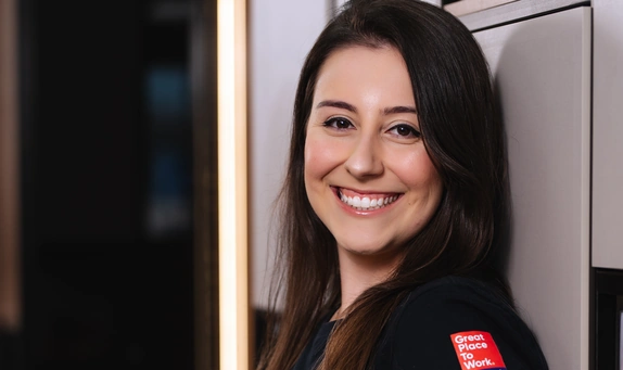 Mulher de cabelos longos e castanhos, sorridente, veste camiseta preta com selo vermelho “Great Place to Work” na manga e está encostada em uma parede, com fundo interno desfocado.