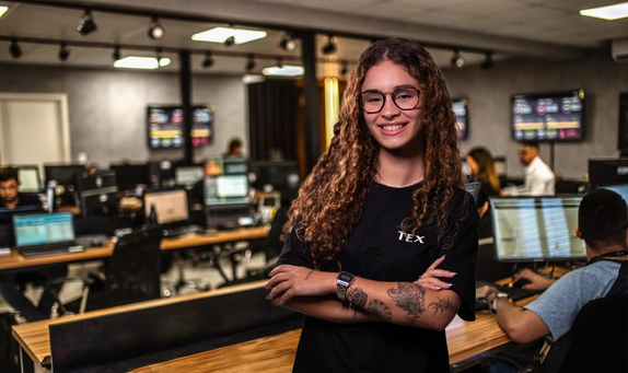 Jovem de cabelo cacheado e óculos sorri com confiança, vestindo uma camiseta preta da TEX, com os braços cruzados, em um escritório moderno com várias pessoas trabalhando em estações de computador ao fundo.