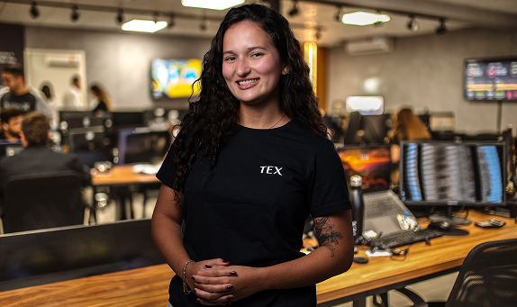Jovem morena de cabelos cacheados sorri para a câmera com mãos cruzadas em frente ao corpo, vestindo uma camiseta preta da TEX, em um ambiente de escritório moderno com persianas ao fundo e tons de luz amarelados.