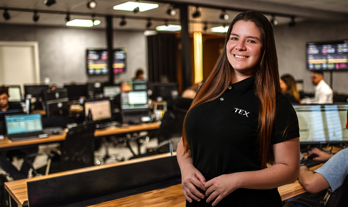 Mulher de cabelos lisos e castanhos, com argola na orelha e piercing no nariz, veste camiseta preta com a palavra "TEX" e posa séria apoiada em uma parede, com fundo desfocado de ambiente interno.