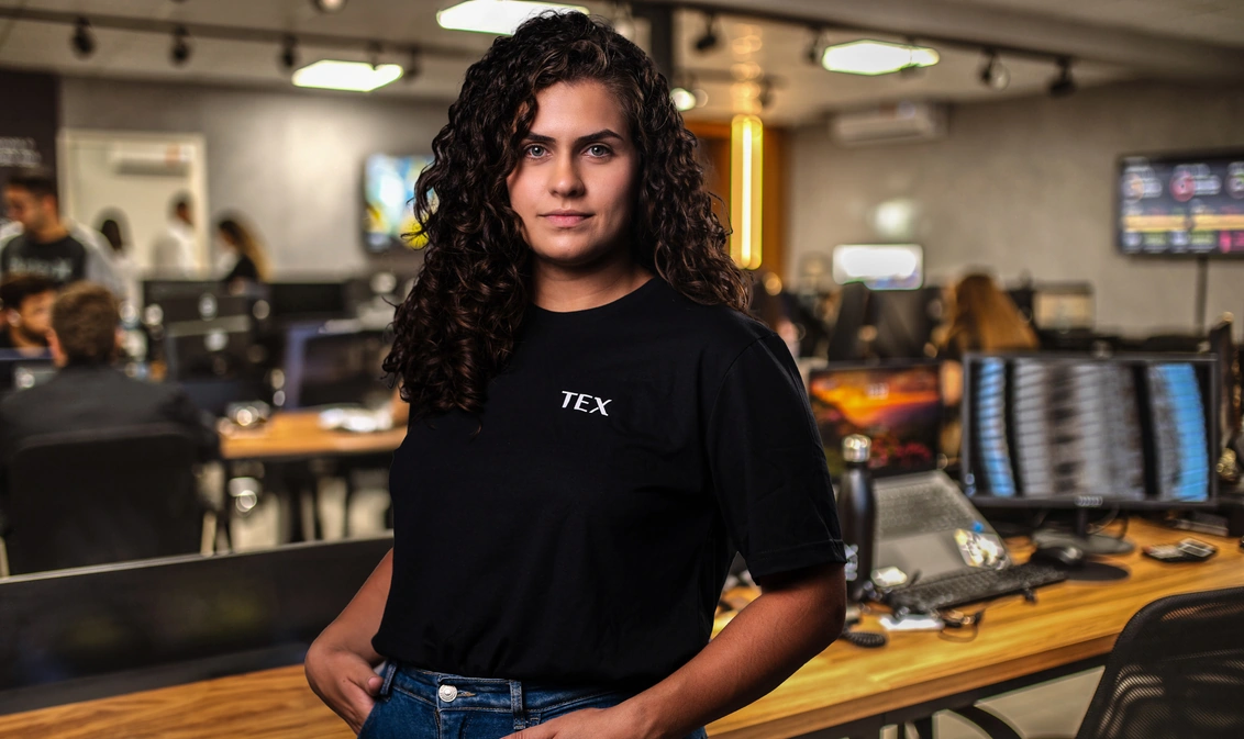 Mulher de cabelos cacheados escuros veste camiseta preta com a palavra "TEX" e posa confiante em um escritório moderno com várias pessoas trabalhando em computadores ao fundo.