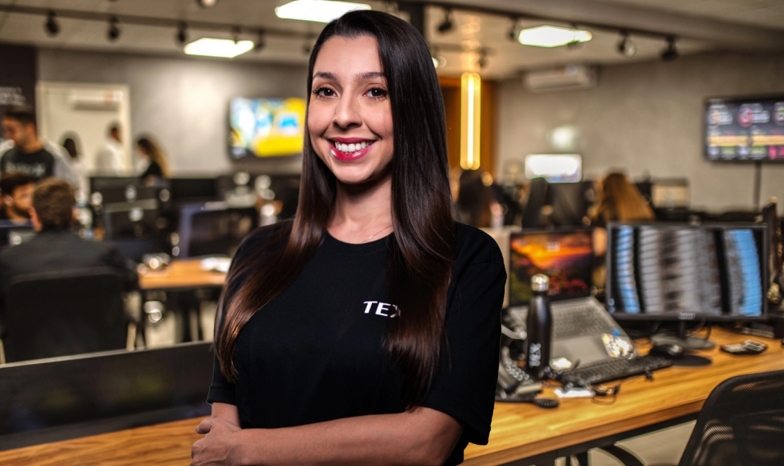 Mulher de cabelos cacheados escuros veste camiseta preta com a palavra "TEX" e posa confiante em um escritório moderno com várias pessoas trabalhando em computadores ao fundo.