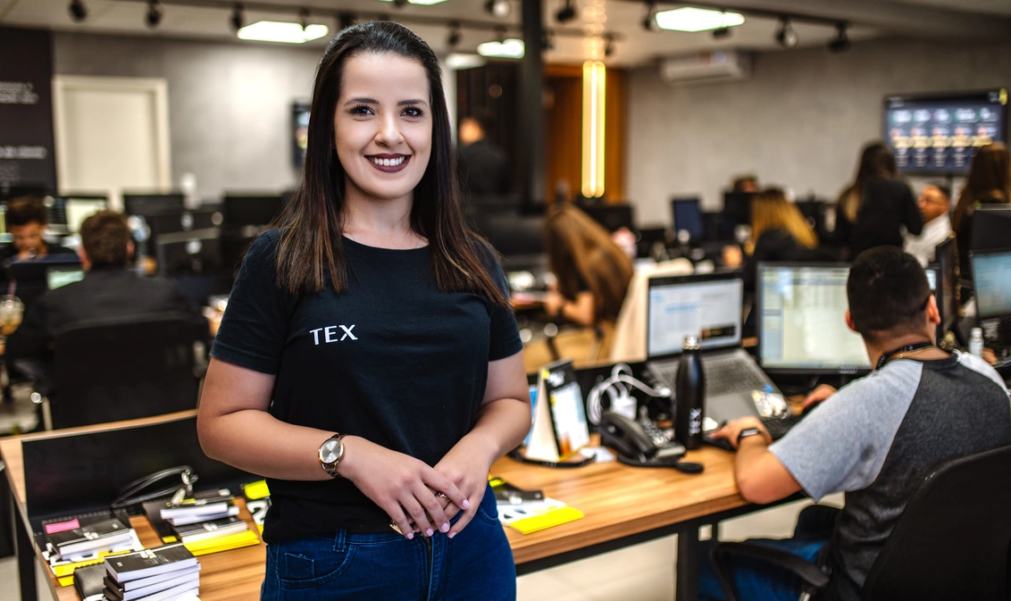 Três pessoas estão de pé e sorriem em um ambiente de escritório, cada uma vestindo roupas com o logotipo da TEX Shipping. Elas estão posicionadas em frente a mesas com computadores, enquanto outras pessoas trabalham ao fundo.